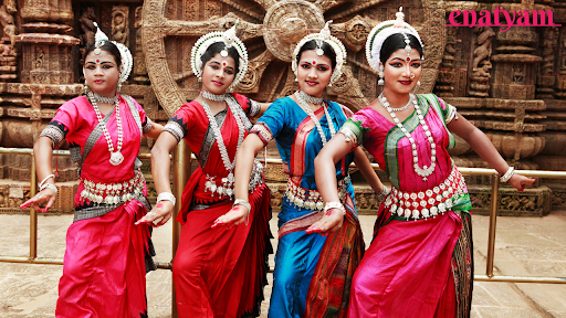 Four dancers in vibrant costumes standing before a stone wheel, showcasing the beauty of the Odissi classical dance form.